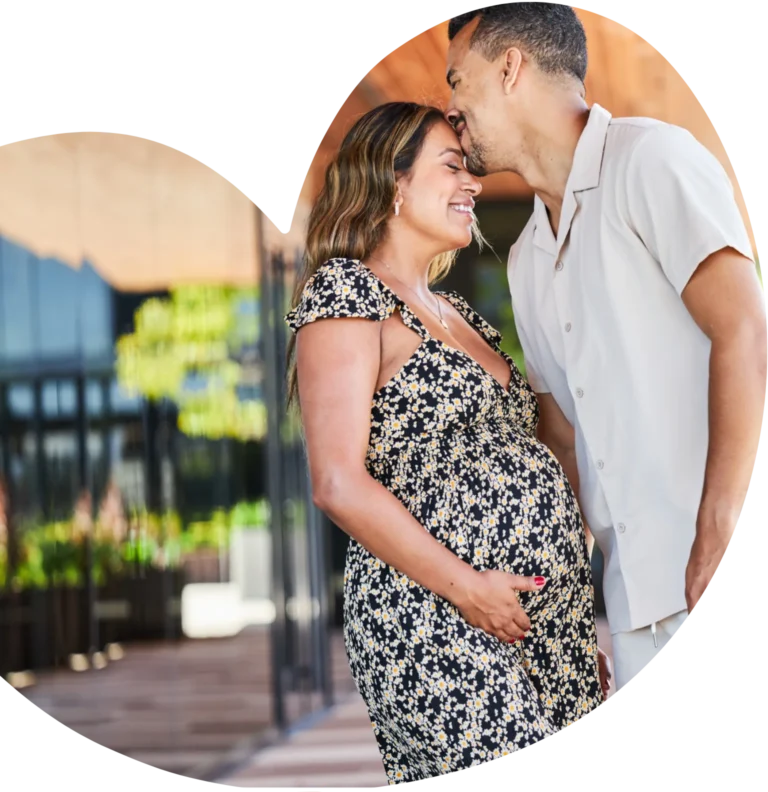 Smiling pregnant woman in a floral dress holding her belly while a man kisses her forehead