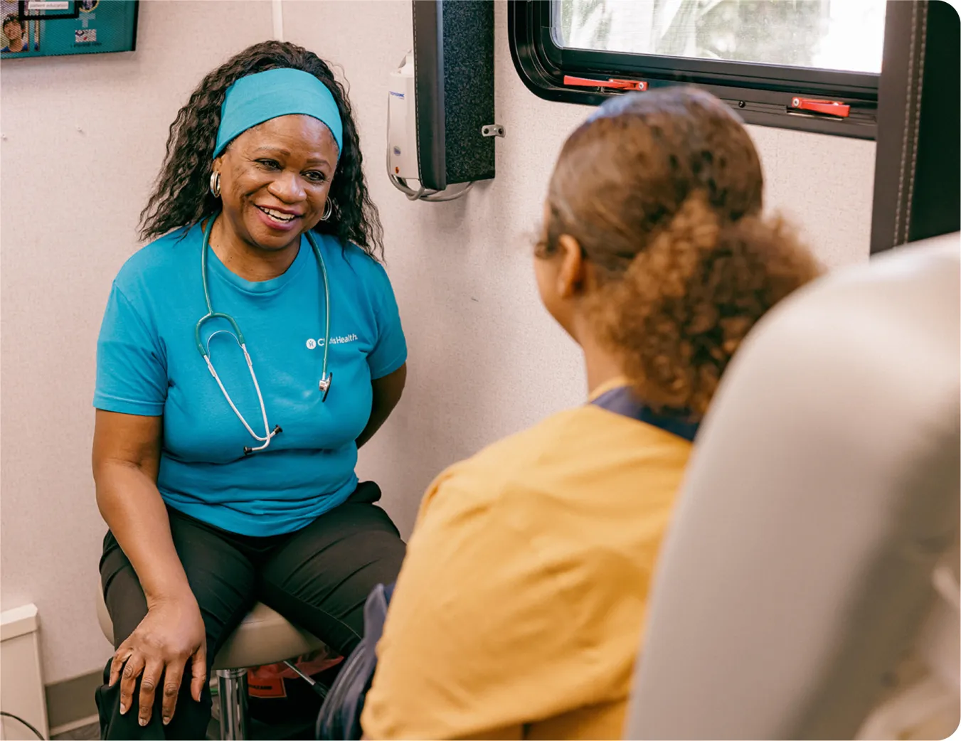 A smiling woman in a Claris Health T-shirt, with a stethoscope, talking with a patient inside the mobile clinic